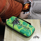A person in an orange shirt and beige pants plays a handheld gaming console while seated. In the foreground, a vibrant green Hyperkin carrying case featuring Cat Noir from "Miraculous: Tales of Ladybug & Cat Noir" is prominently displayed, showcasing the character's image and name.