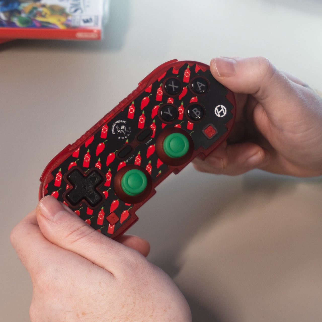 Person holding a red and black gaming controller with green thumbsticks and X, Y, B, A buttons, featuring a chili-themed design and a video game case in the background.