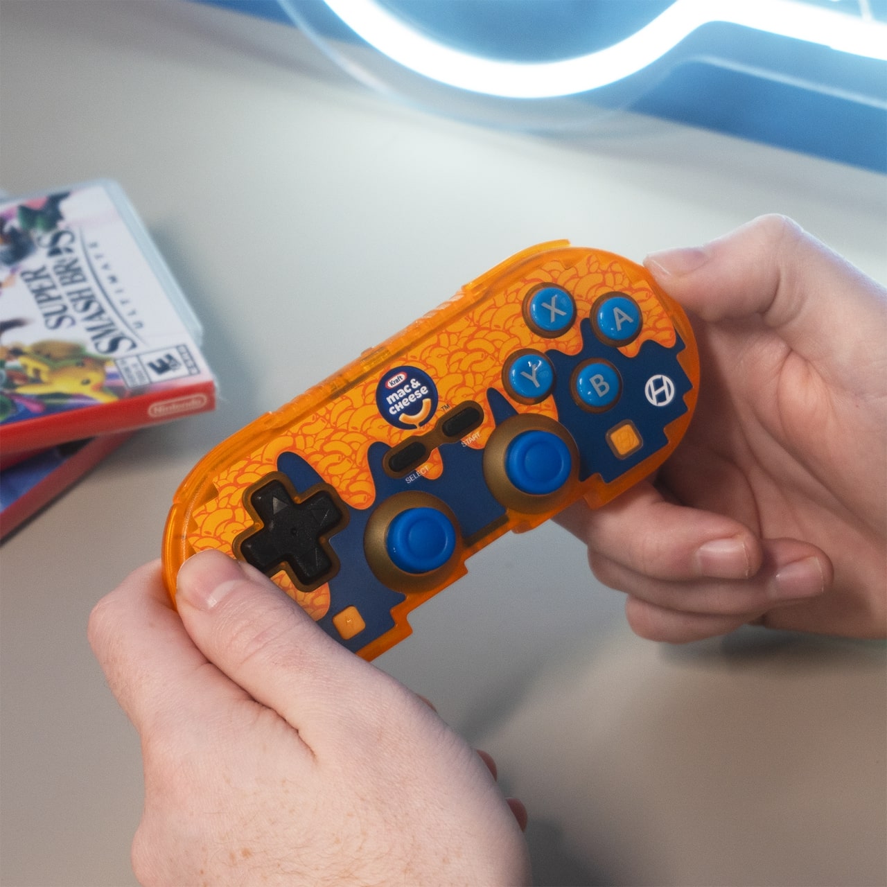 Person holding orange and blue Hyperkin gaming controller with Kraft Mac & Cheese design, blue buttons, and Nintendo Switch game cases in the background.
