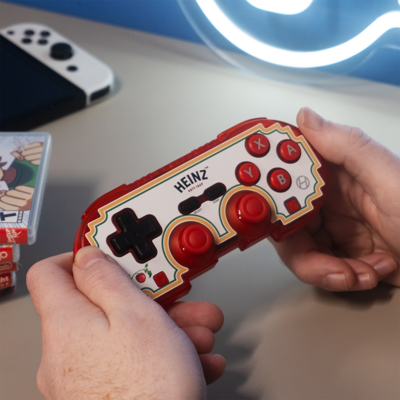 Custom Heinz-themed Hyperkin Bluetooth gaming controller in red and white, featuring ABXY buttons, dual red joysticks, and central Heinz logo. Held by a person with a Nintendo Switch console and game case visible in the background.