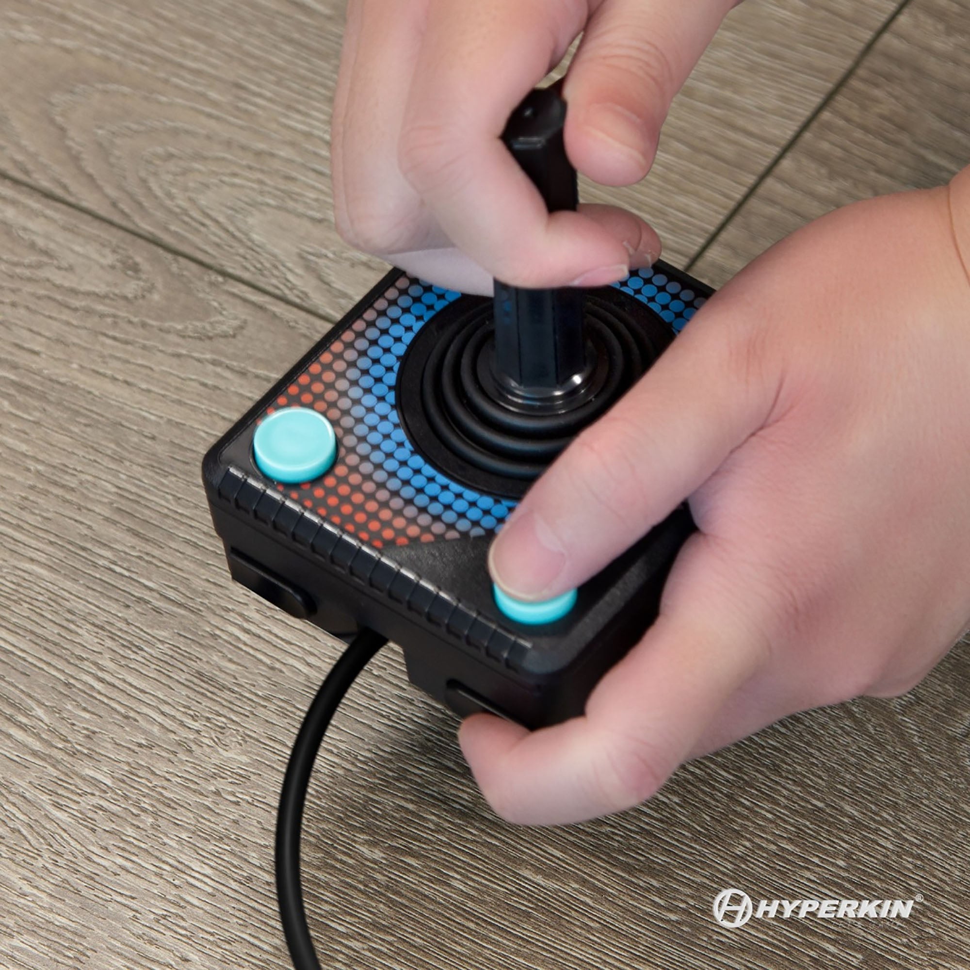 Close-up of hands using retro-style black Hyperkin joystick controller with blue buttons and red-blue pixelated base design; cable connected, set against wooden floor background with Hyperkin branding.