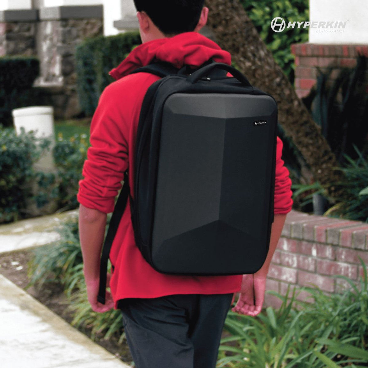 Person wearing red hoodie walking outdoors with large black Hyperkin geometric-shaped backpack. Sleek design shown against greenery and brick wall background.