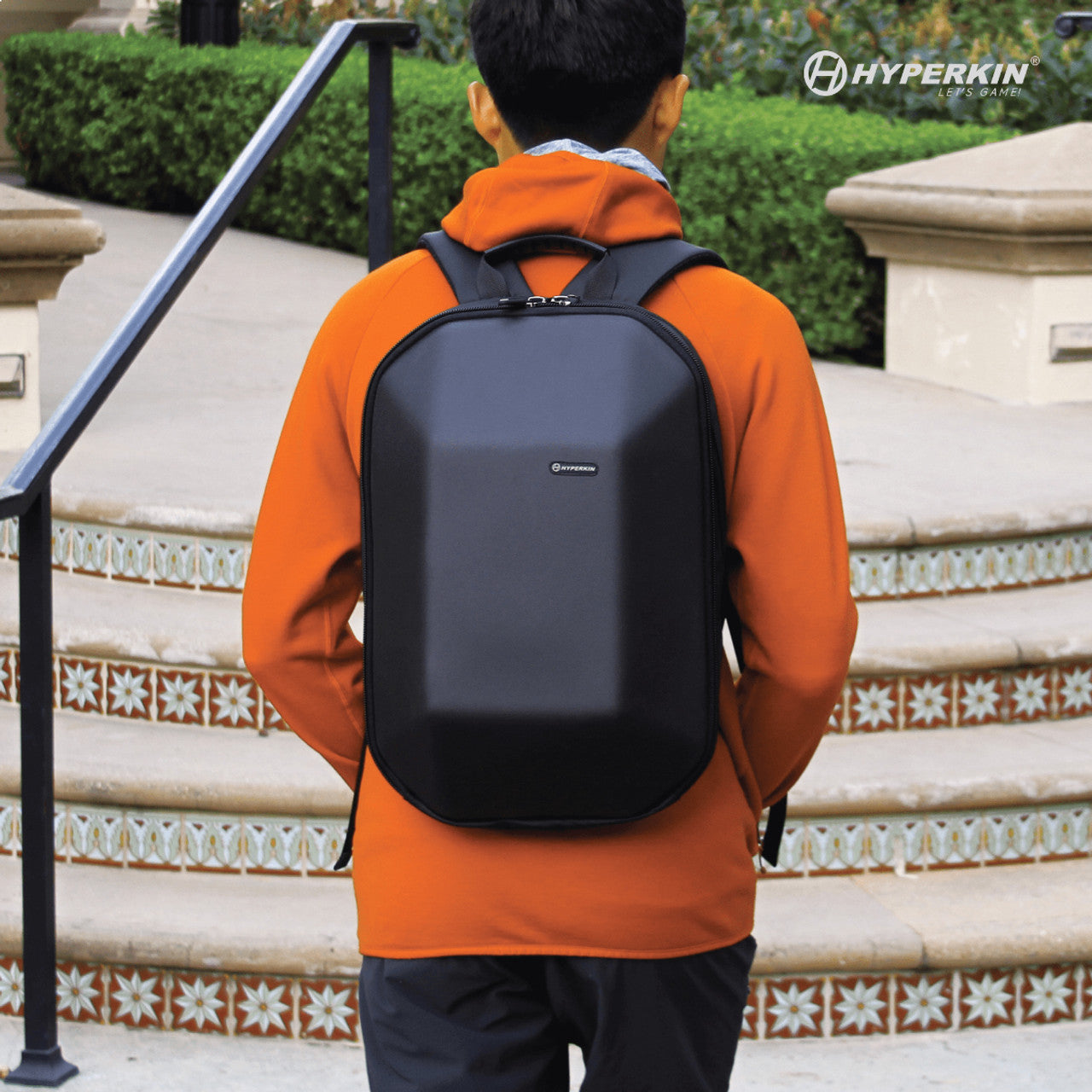 Person wearing orange jacket displaying Hyperkin black backpack outdoors near tiled steps and greenery. Logo visible in top right corner.